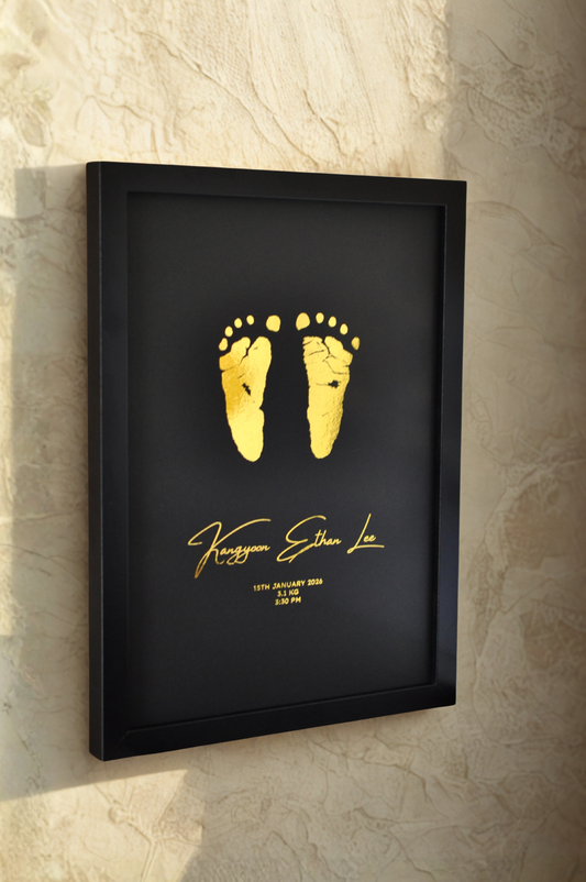Framed gold baby footprints on a black background with personalized text, mounted on a textured wall.