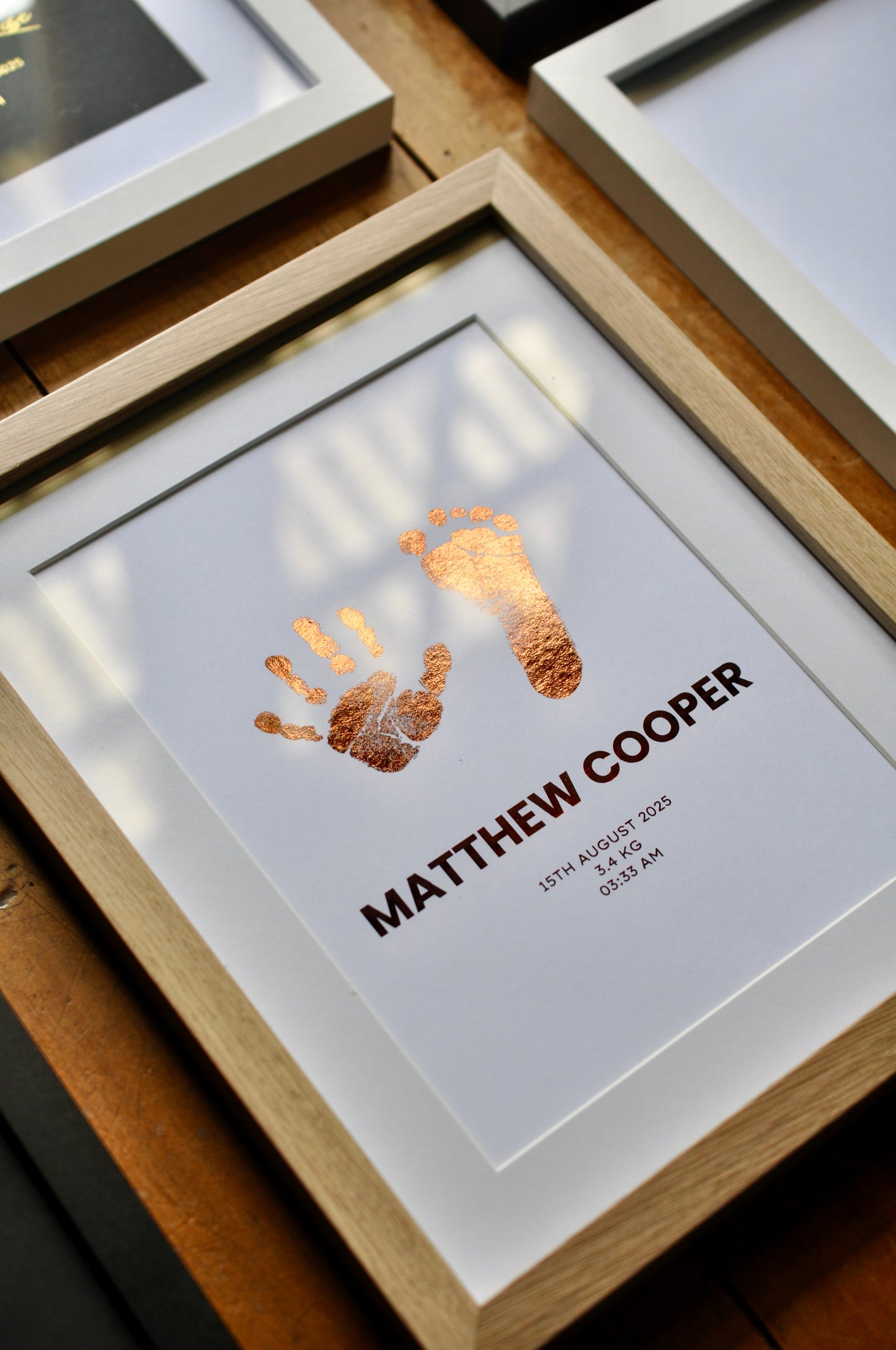 Framed print with handprint and footprint in rose foil and name on a wooden surface.