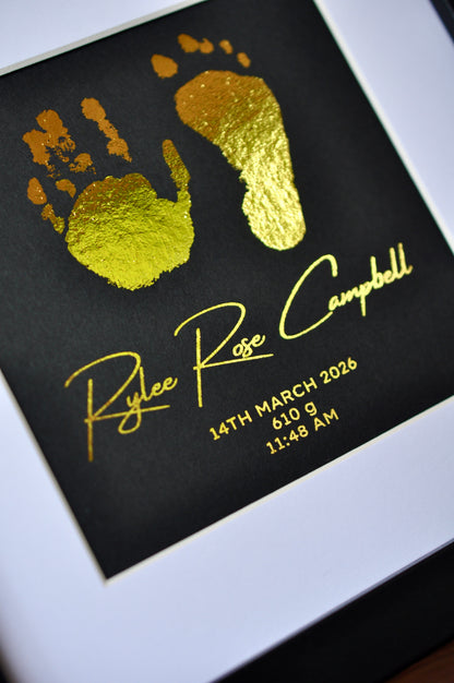Gold hand and footprints on a black background with text, framed.