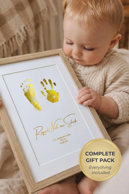Baby holding a framed handprint and footprint with gold foil print, sitting on a soft surface.