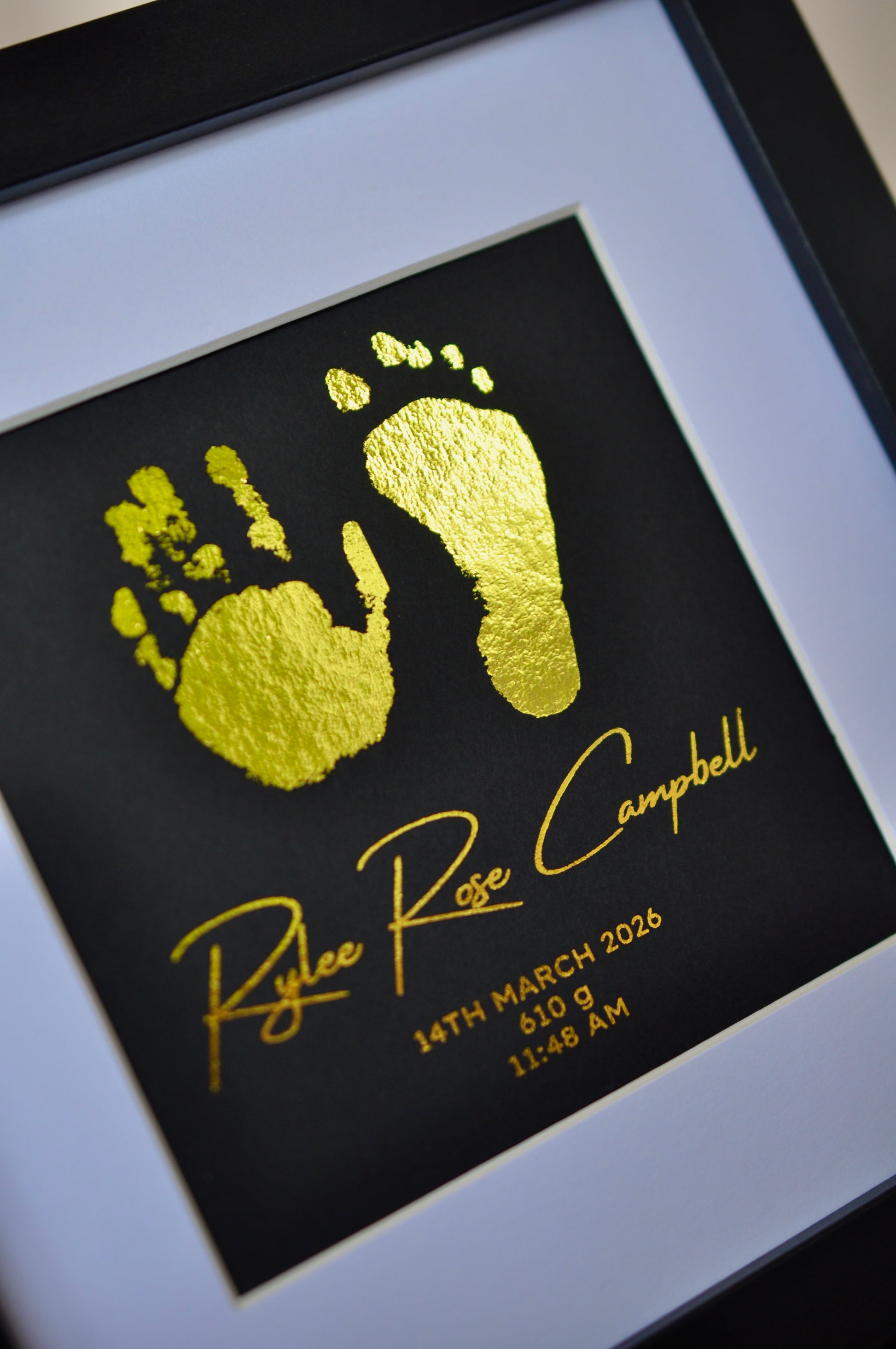 Framed artwork with gold baby footprints and name on black background