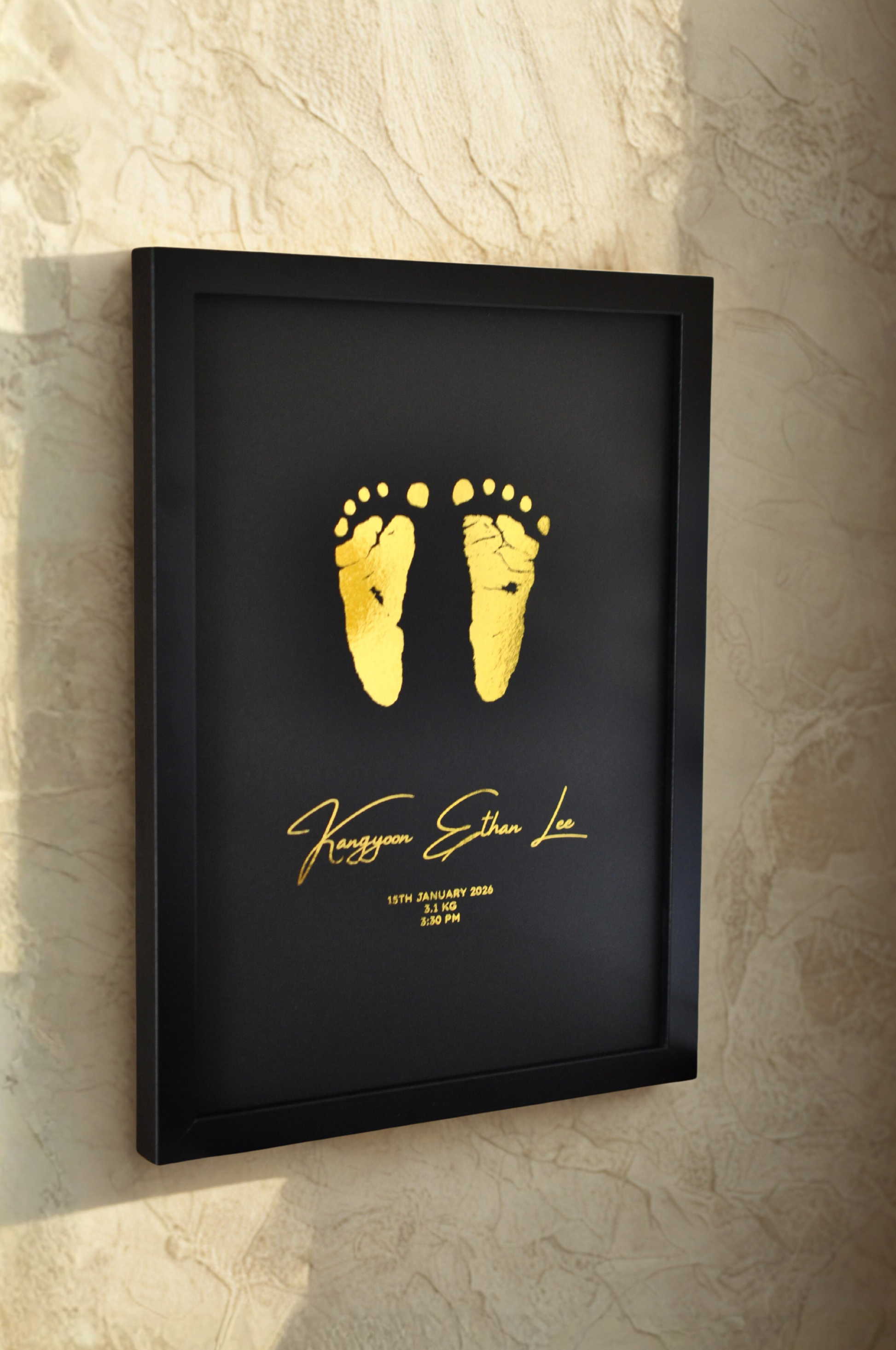 Framed gold baby footprints on a black background with personalized text, mounted on a textured wall.