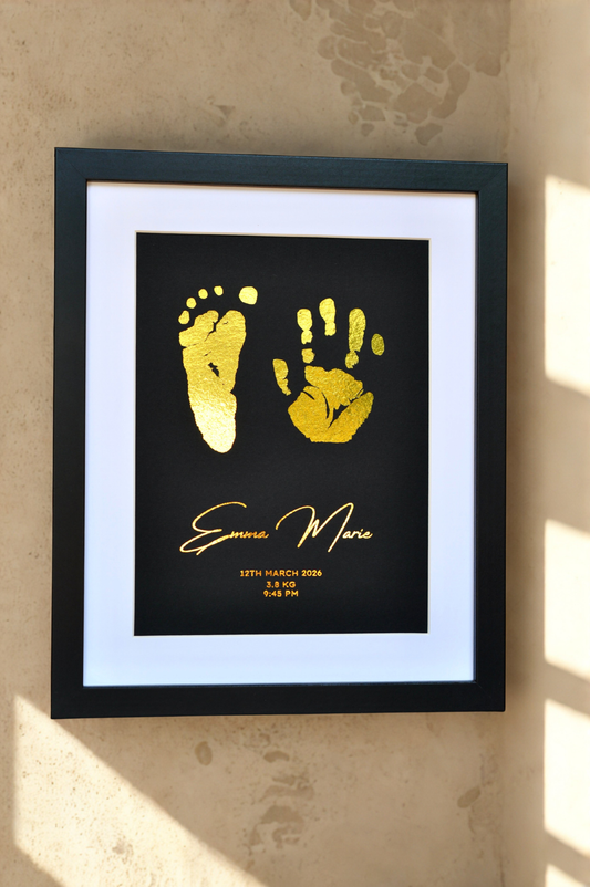 Framed gold hand and footprints on a black background with text, mounted on a beige wall.